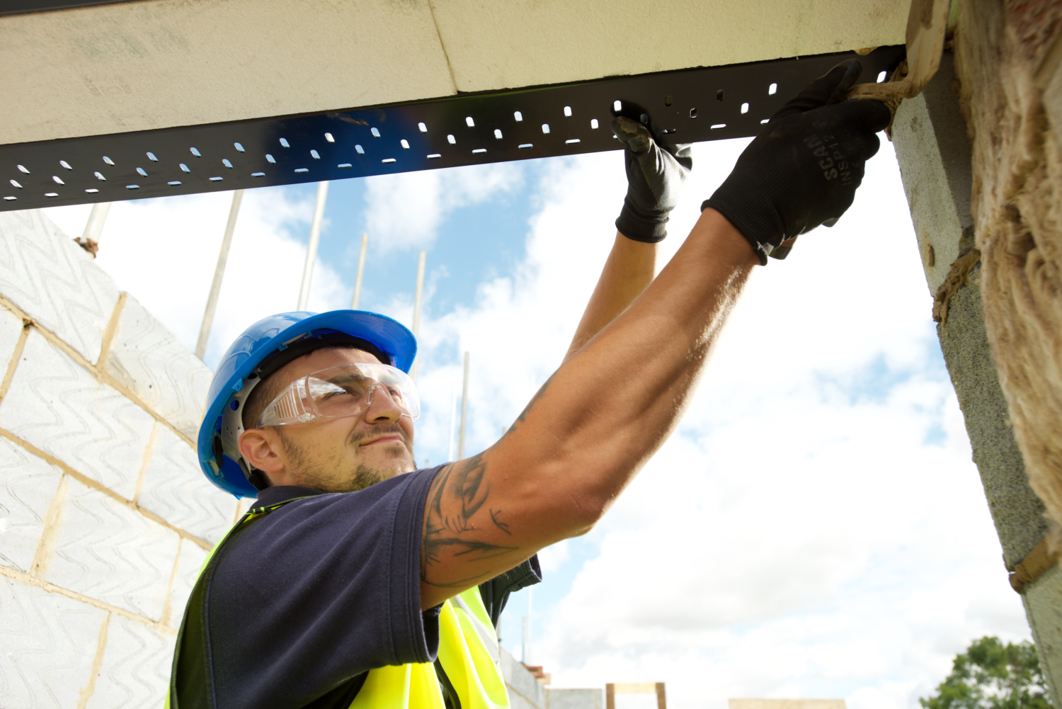Construction worker securing a steel lintel wearing a hard hat and safety goggles. He stands between unfinished brick walls under a clear blue sky.