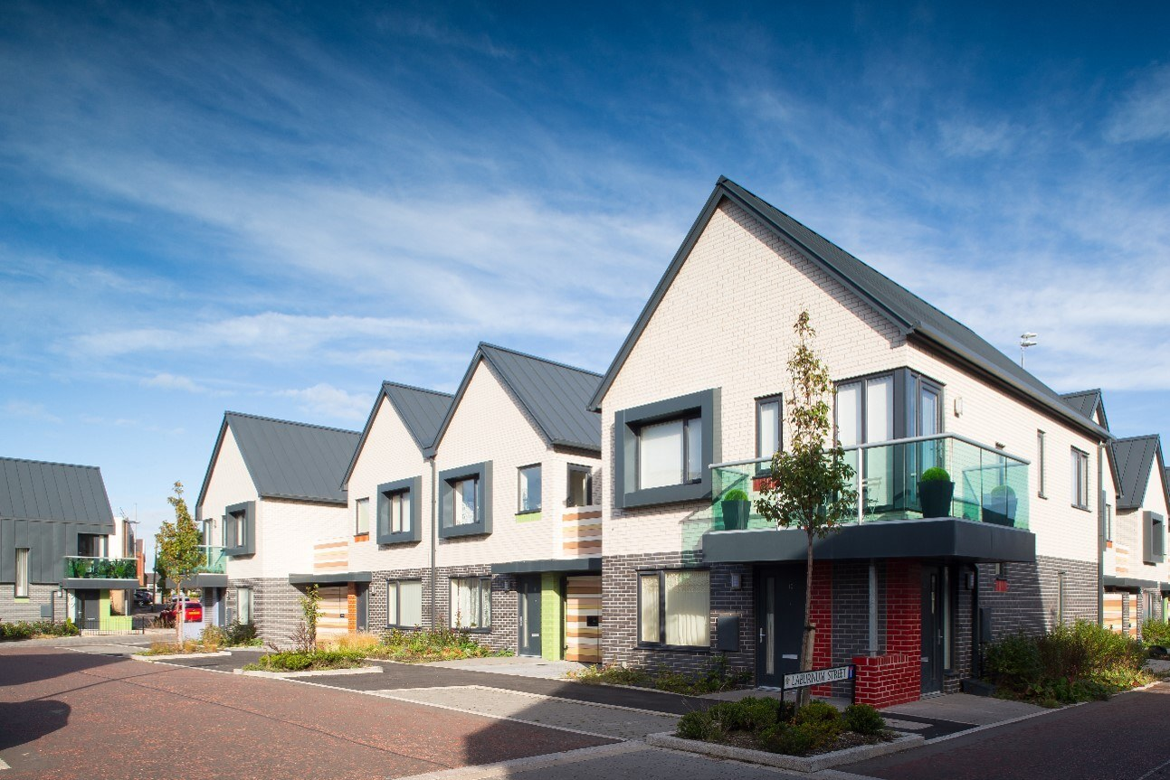 Row of modern, two-story houses with standing seam roofs and balconies, situated along a quiet street lined with young trees and landscaped gardens