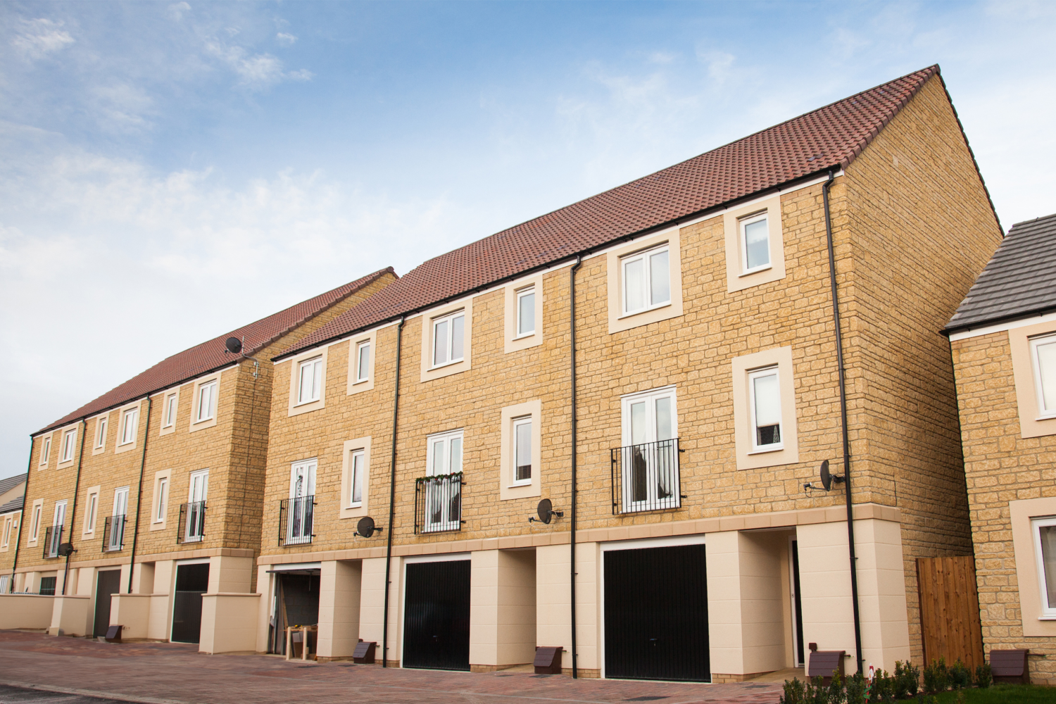 Row of modern, three-story townhouses with stone facades and red-tiled roofs sit under a blue sky. Each unit has Catnic juliet balconies, large windows, and black garage doors.