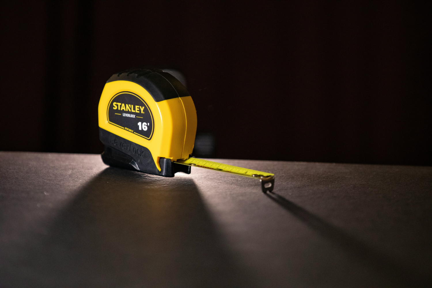 A yellow Stanley Leverlock tape measure extends its metal measuring tape across a dark surface, casting a shadow in a dimly lit environment. Text: "STANLEY LEVERLOCK 16' LEVELLOCK."