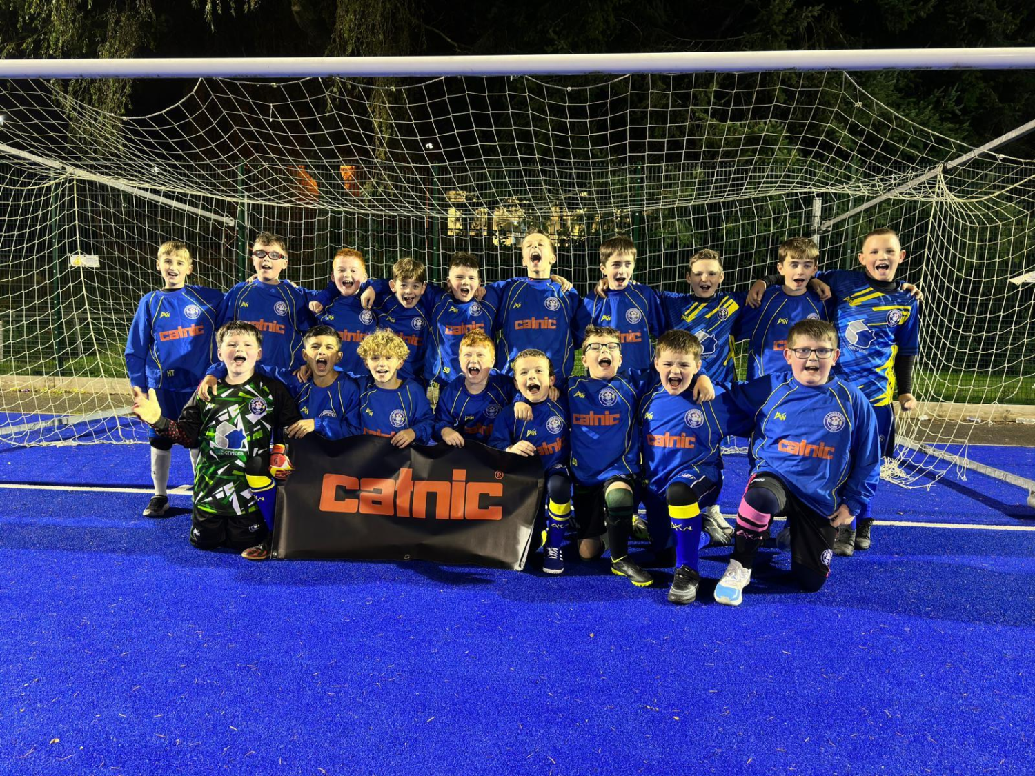 A group of young soccer players wearing blue uniforms pose excitedly in front of a goal. They hold a banner with "catnic" text on a vibrant blue field, surrounded by trees.